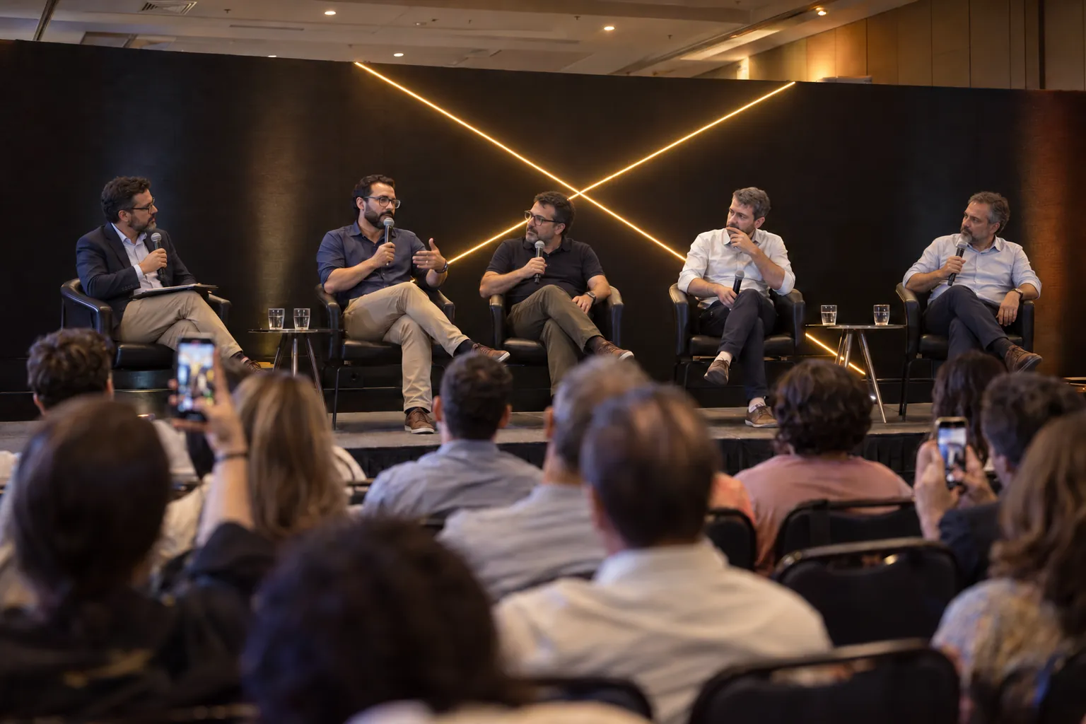 Painel mediado com especialistas no palco, diante da plateia, com o X de luz amarela ao fundo